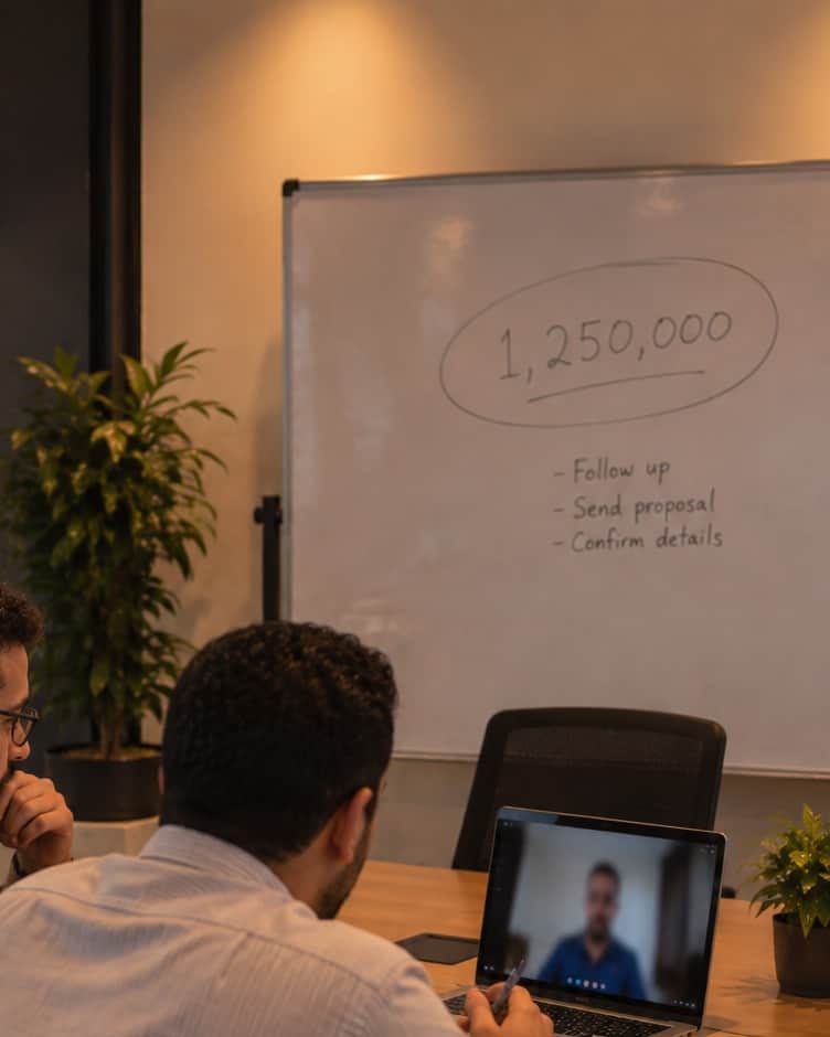 Two founders on a video call with a client, a single revenue figure circled on the whiteboard behind them